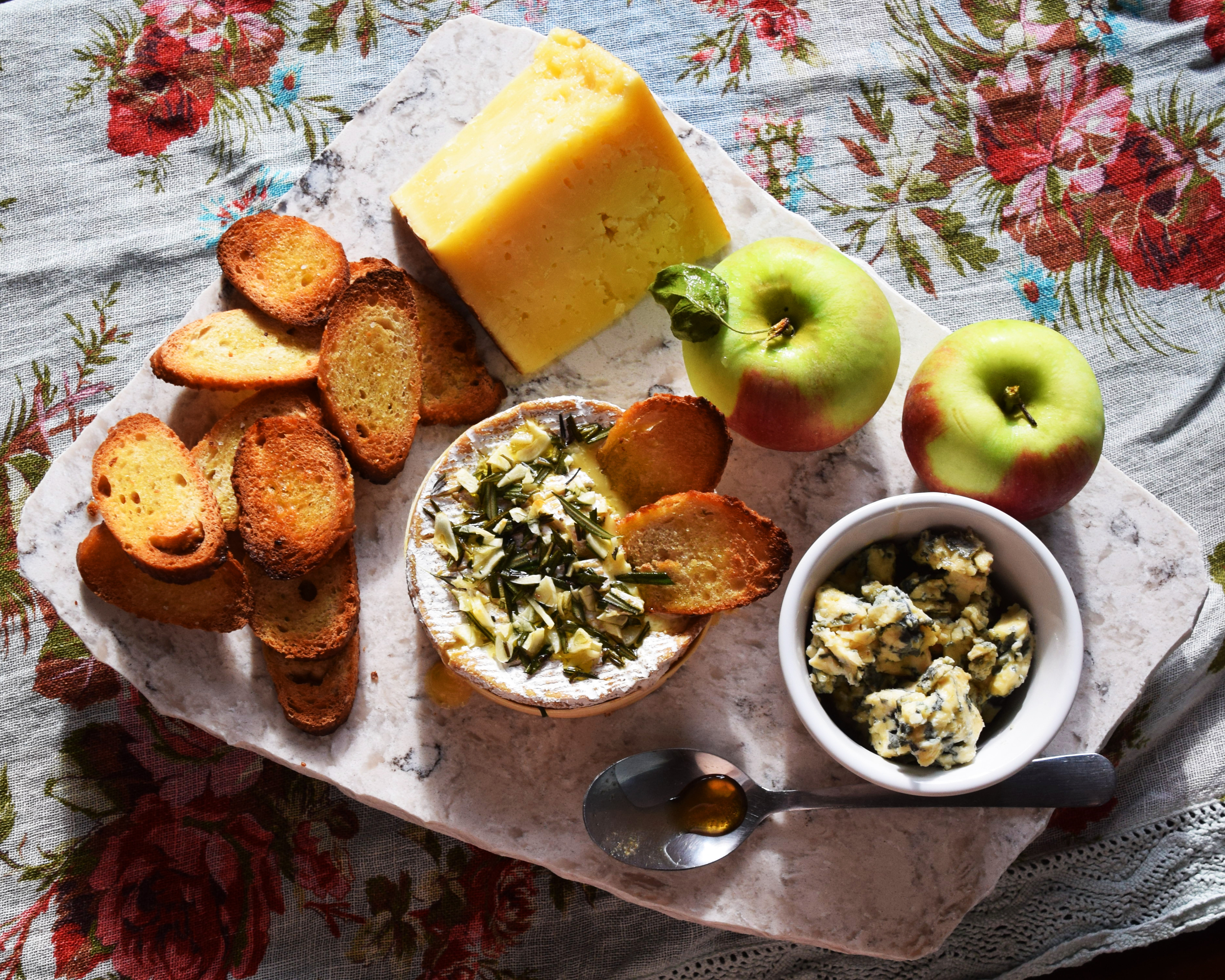 3 cheeses on a stone board surrounded by apples, a spoon, and toasts